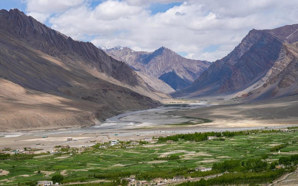 Green fields of a valley village with a winding river and snow-streaked mountains under a cloudy sky.