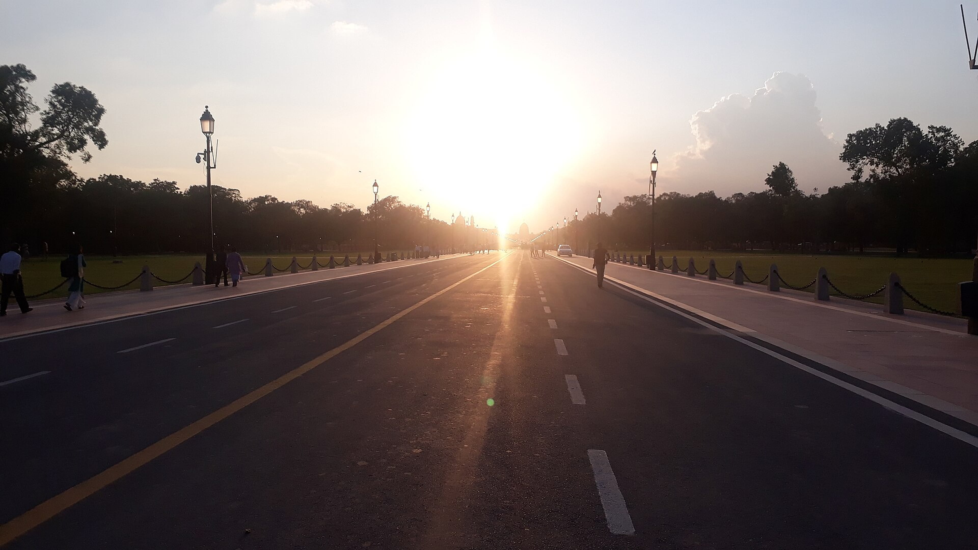 Evening sun over Kartavya Path in New Delhi, with lamp posts lining the road and a few pedestrians on the footpath.