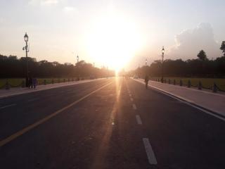 Evening sun over Kartavya Path in New Delhi, with lamp posts lining the road and a few pedestrians on the footpath.
