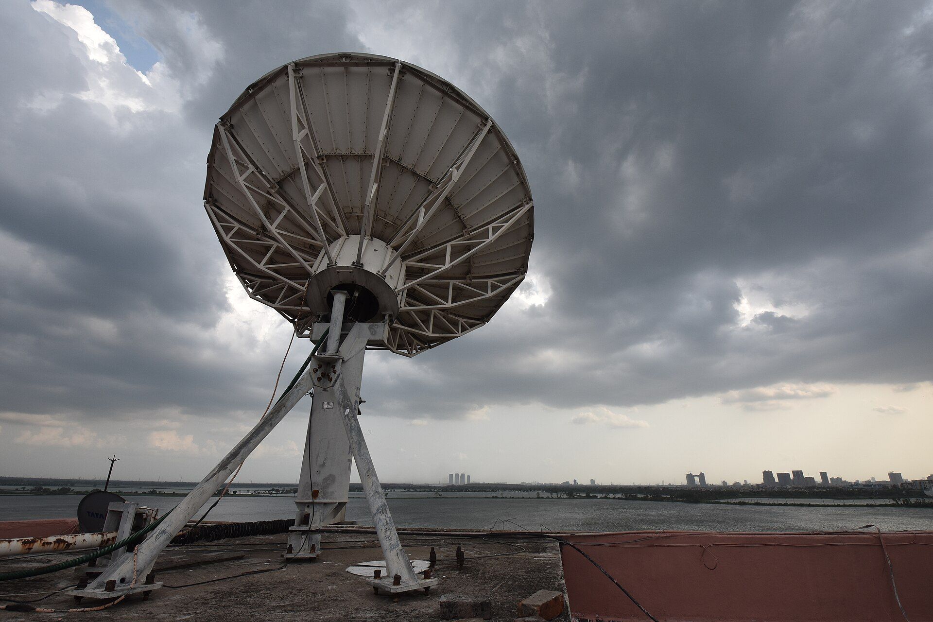 Large satellite dish on a rooftop under dark storm clouds, with water and a distant city skyline behind it