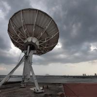 Large satellite dish on a rooftop under dark storm clouds, with water and a distant city skyline behind it