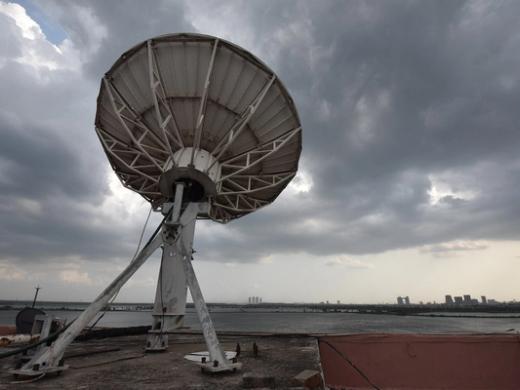 Large satellite dish on a rooftop under dark storm clouds, with water and a distant city skyline behind it