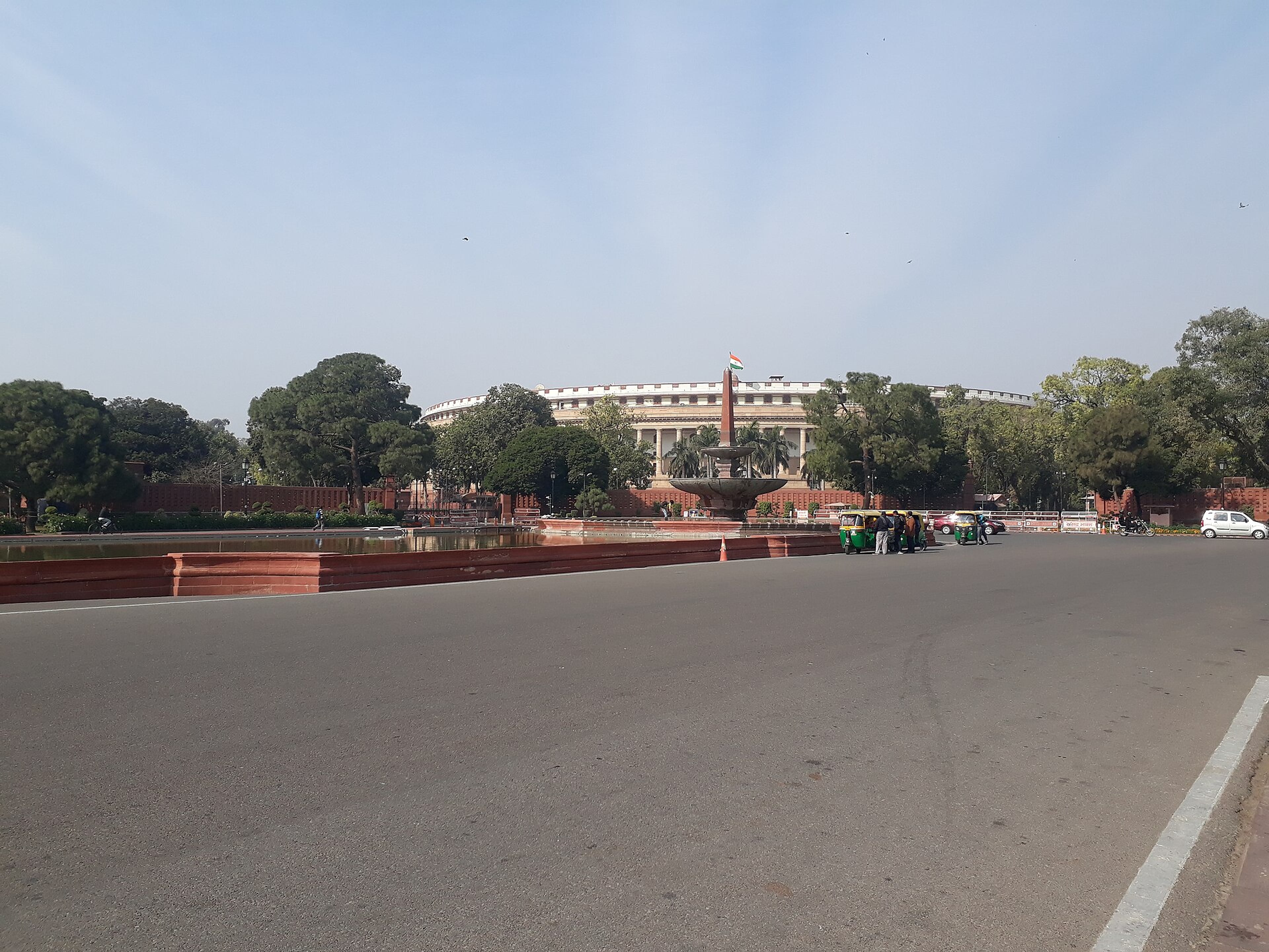 Wide road leading to a fountain with an obelisk, a curved white building behind trees, and an auto-rickshaw.