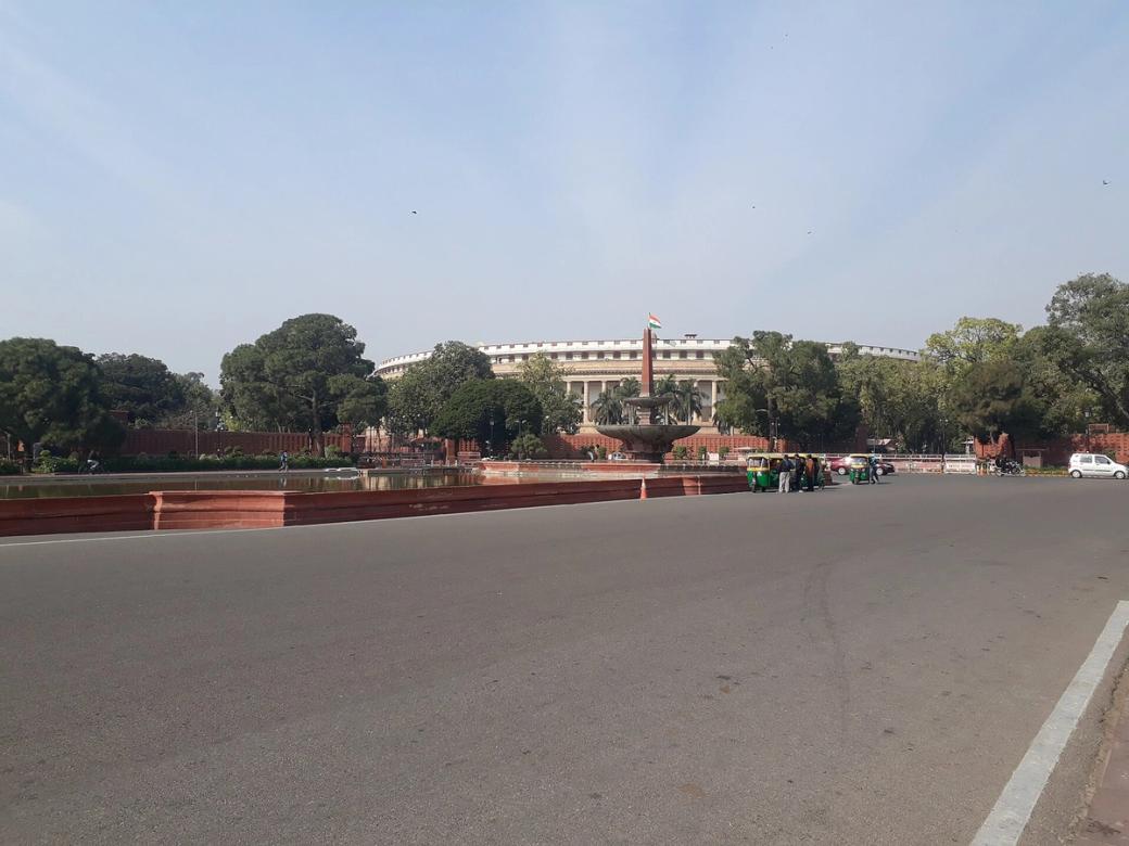 Wide road leading to a fountain with an obelisk, a curved white building behind trees, and an auto-rickshaw.