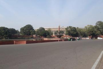 Wide road leading to a fountain with an obelisk, a curved white building behind trees, and an auto-rickshaw.