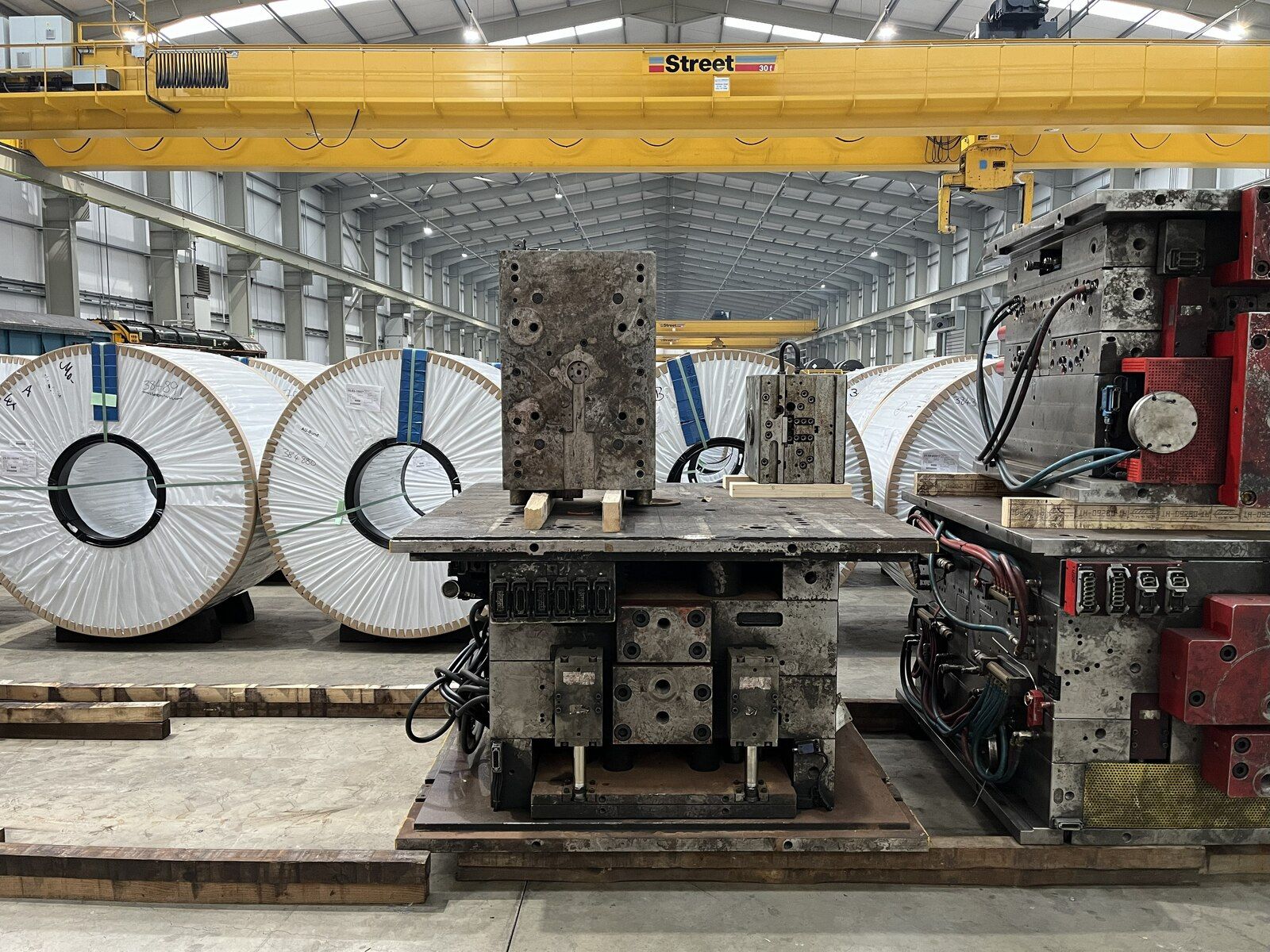 Large white-wrapped steel coils and metal industrial machinery on a factory floor under a yellow overhead crane