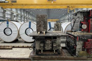 Large white-wrapped steel coils and metal industrial machinery on a factory floor under a yellow overhead crane