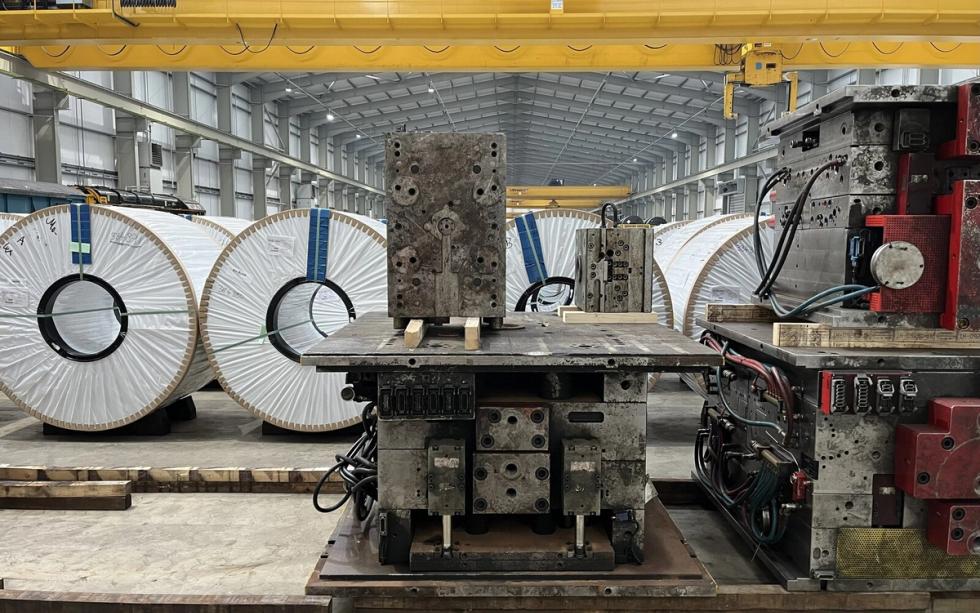 Large white-wrapped steel coils and metal industrial machinery on a factory floor under a yellow overhead crane