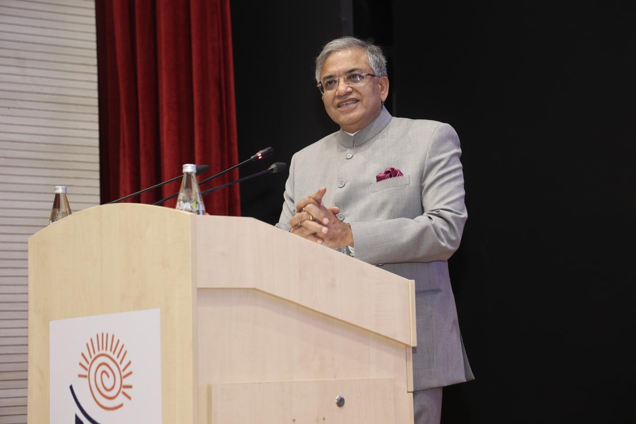 A man in a grey bandhgala jacket and glasses speaks at a wooden podium with microphones, red curtain behind him