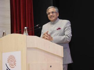 A man in a grey bandhgala jacket and glasses speaks at a wooden podium with microphones, red curtain behind him