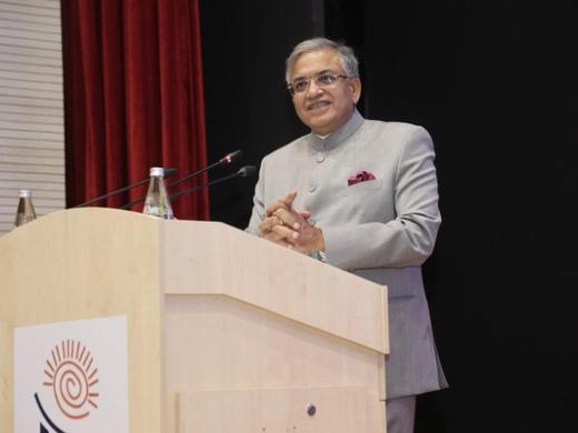 A man in a grey bandhgala jacket and glasses speaks at a wooden podium with microphones, red curtain behind him