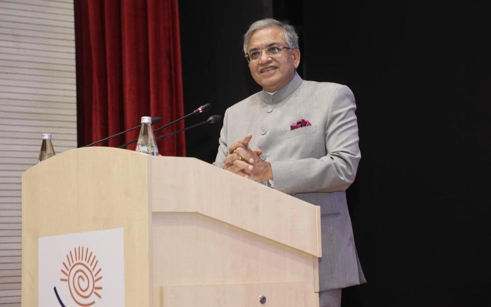 A man in a grey bandhgala jacket and glasses speaks at a wooden podium with microphones, red curtain behind him