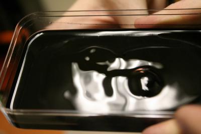 Dark ferrofluid blob in a clear tray, with fingers holding the tray at the edges
