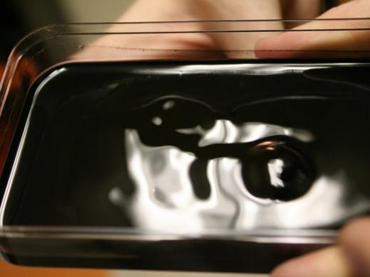 Dark ferrofluid blob in a clear tray, with fingers holding the tray at the edges