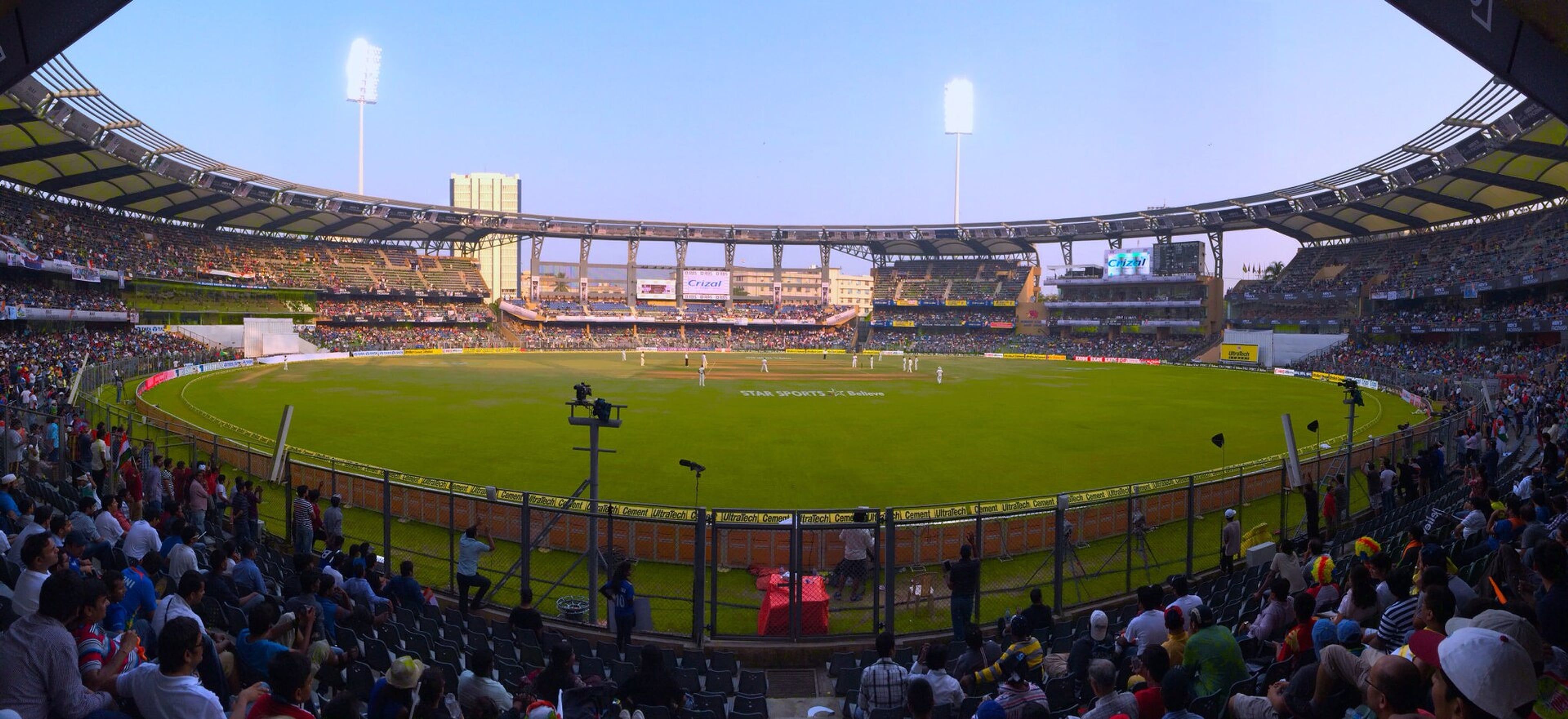 Panoramic view of Wankhede Stadium with crowded stands and players on the pitch in the evening light.