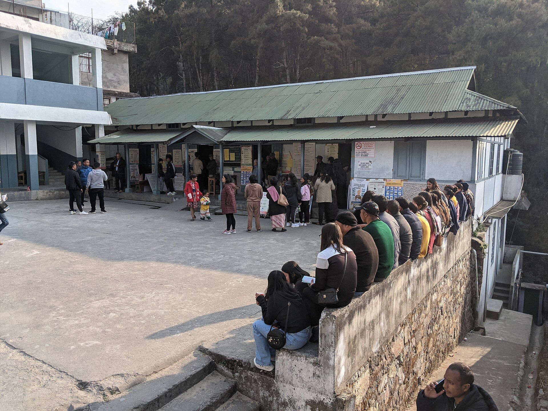 People queue outside a polling station with corrugated roofs, some waiting on a low wall, in Shillong, Meghalaya.