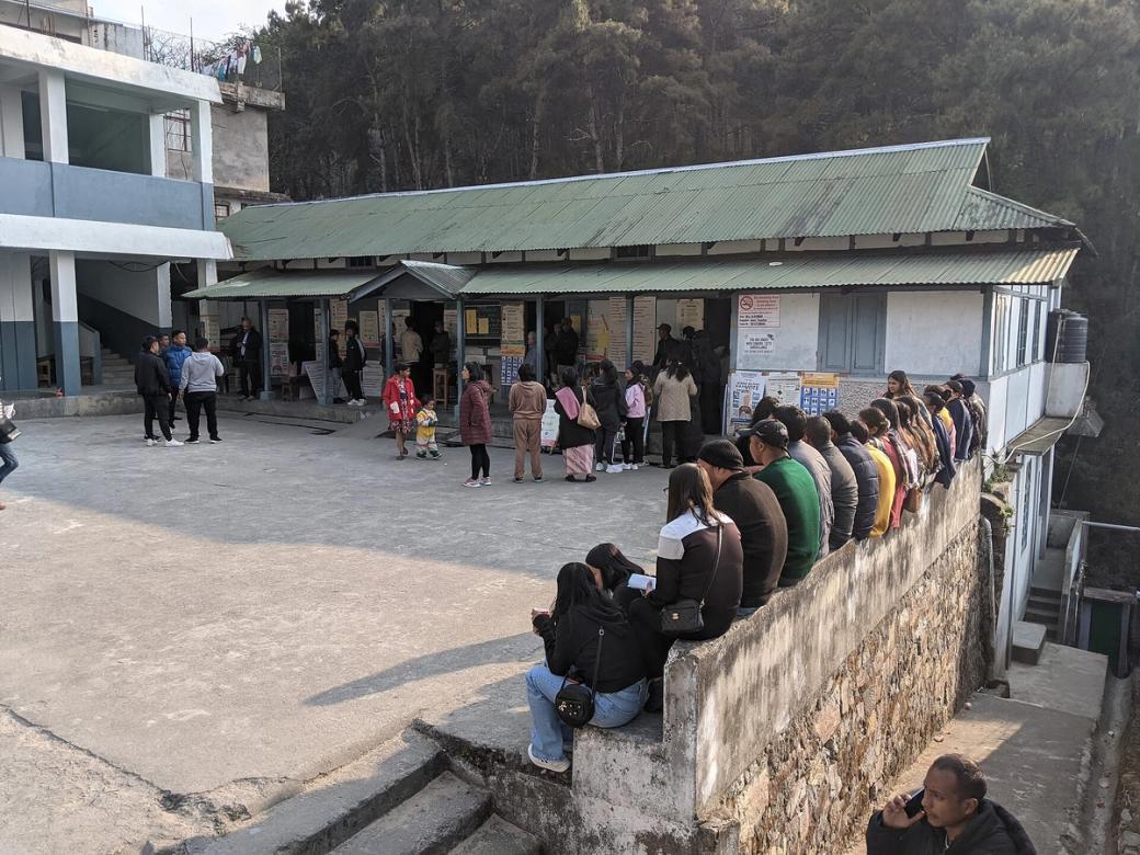 People queue outside a polling station with corrugated roofs, some waiting on a low wall, in Shillong, Meghalaya.