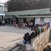 People queue outside a polling station with corrugated roofs, some waiting on a low wall, in Shillong, Meghalaya.