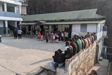 People queue outside a polling station with corrugated roofs, some waiting on a low wall, in Shillong, Meghalaya.