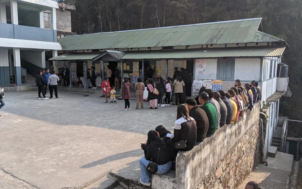 People queue outside a polling station with corrugated roofs, some waiting on a low wall, in Shillong, Meghalaya.