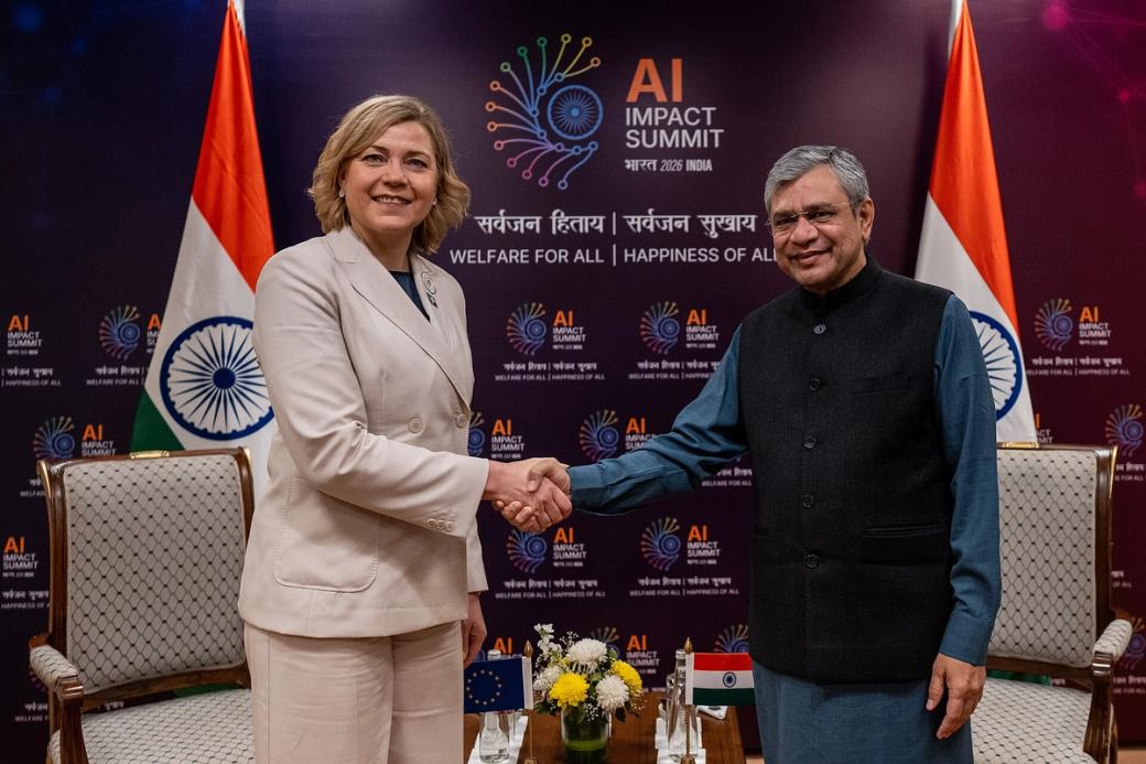Ashwini Vaishnaw and Henna Virkkunen shake hands before an AI Impact Summit backdrop with Indian and EU flags.