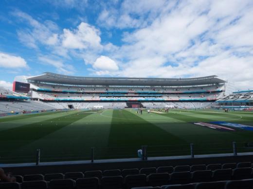 Empty Eden Park stadium in daylight, with a few players warming up on the pitch under a blue sky.