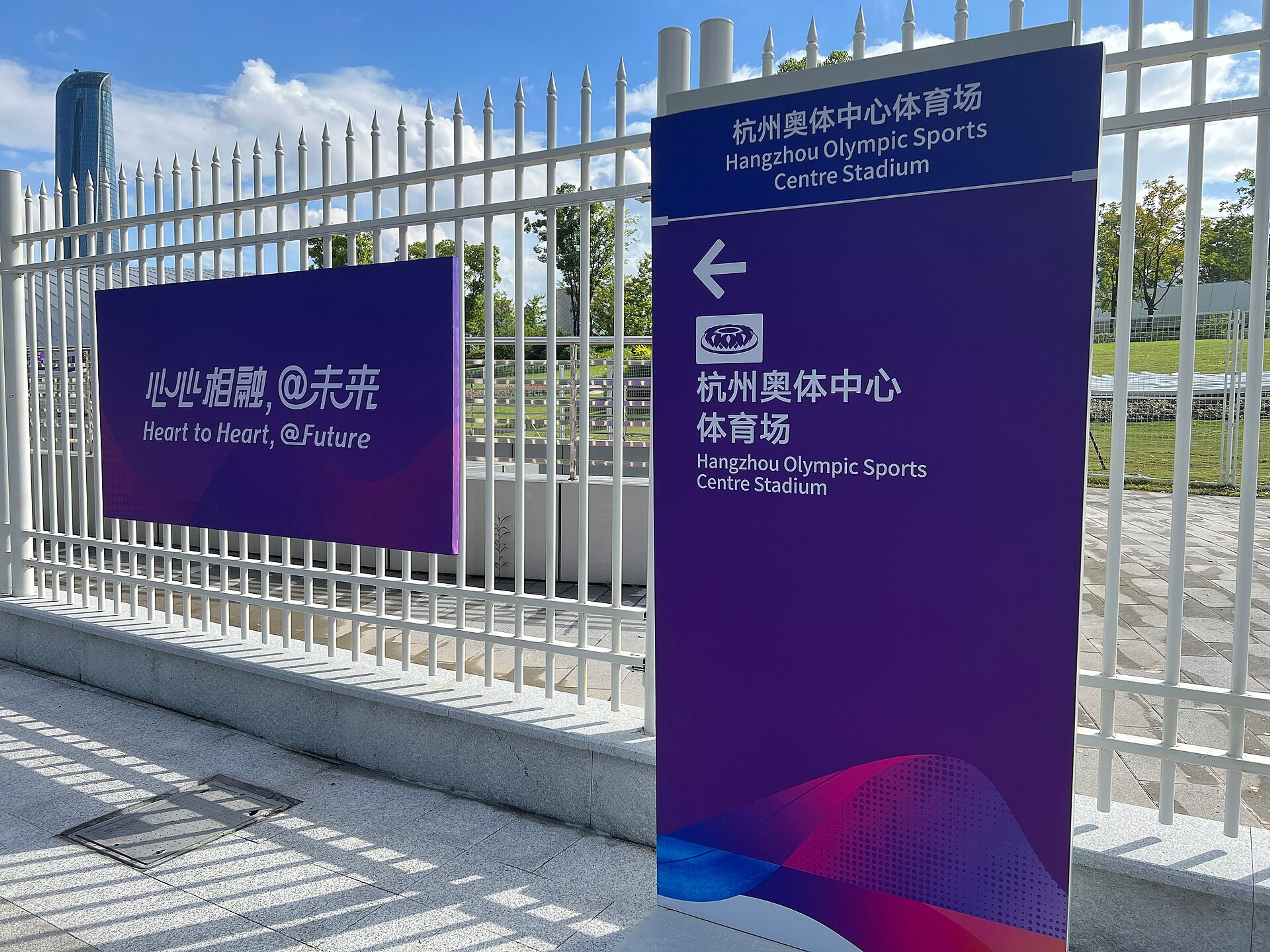 Asian Games slogan sign and Hangzhou Olympic Sports Centre stadium direction sign beside a white fence.