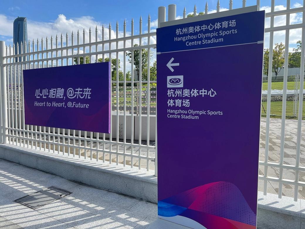 Asian Games slogan sign and Hangzhou Olympic Sports Centre stadium direction sign beside a white fence.