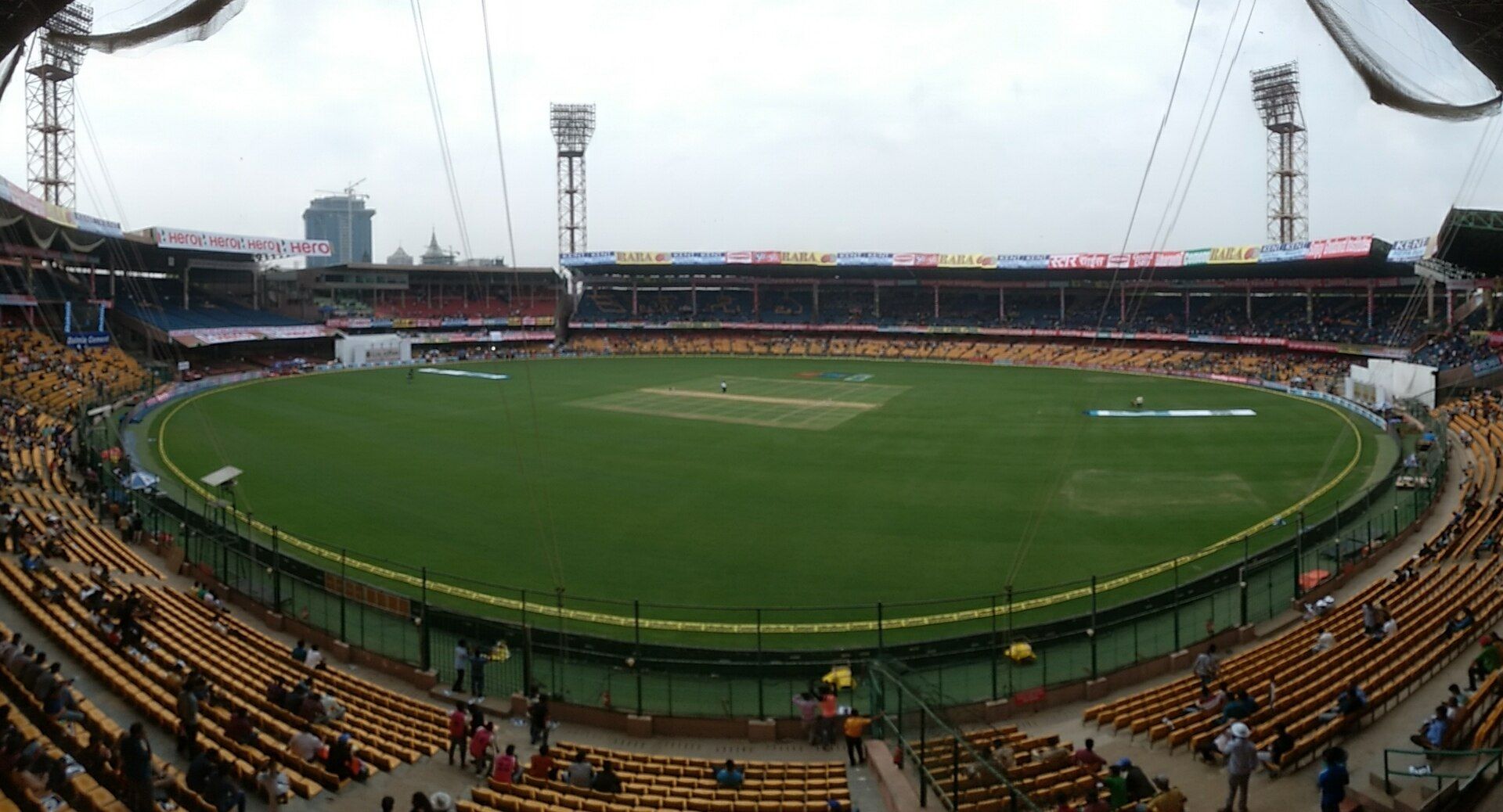 Panoramic view of a cricket stadium in Bengaluru with yellow seating, a green field and floodlight towers