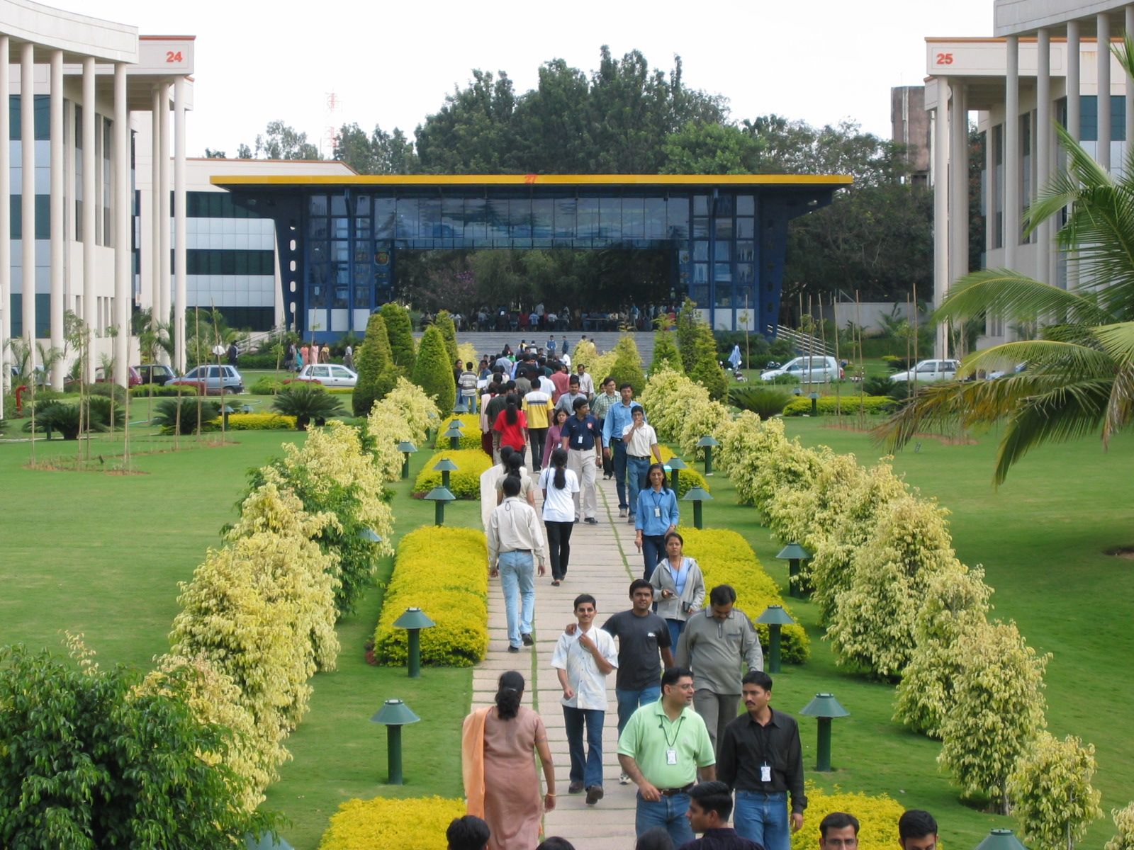 Employees with ID badges walk along a paved garden path between office buildings on a green campus