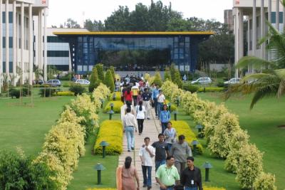 Employees with ID badges walk along a paved garden path between office buildings on a green campus