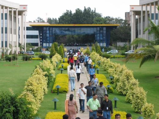 Employees with ID badges walk along a paved garden path between office buildings on a green campus