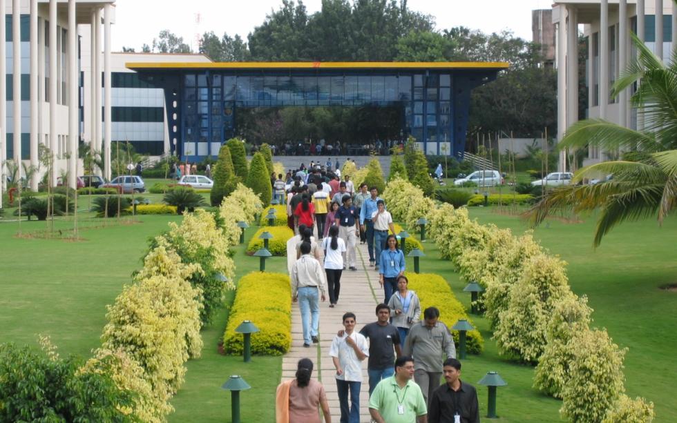 Employees with ID badges walk along a paved garden path between office buildings on a green campus