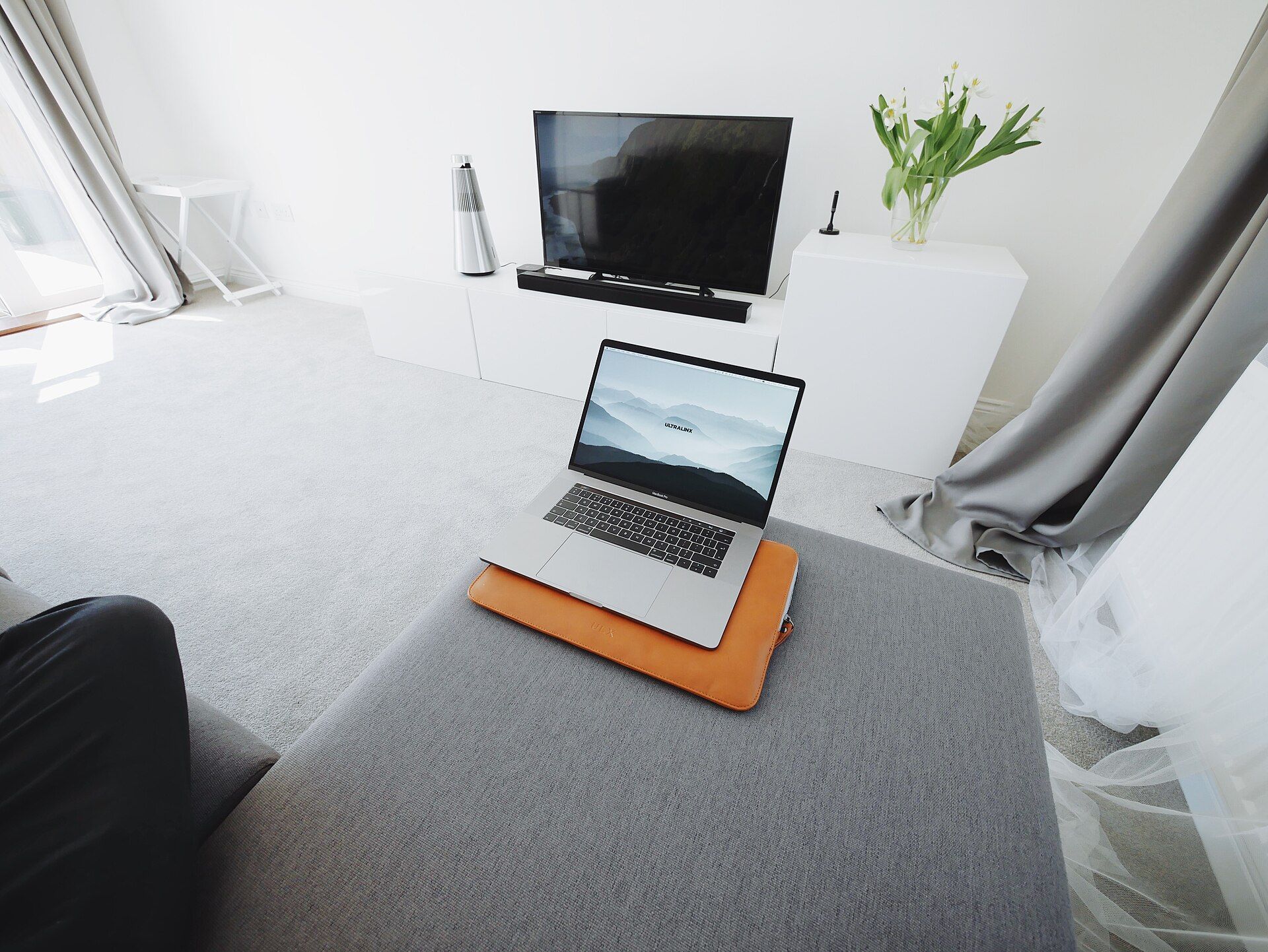 Open laptop on a grey ottoman in a bright living room with a wall-mounted flat-screen TV and a white cabinet.