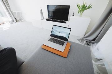 Open laptop on a grey ottoman in a bright living room with a wall-mounted flat-screen TV and a white cabinet.