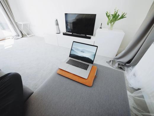 Open laptop on a grey ottoman in a bright living room with a wall-mounted flat-screen TV and a white cabinet.
