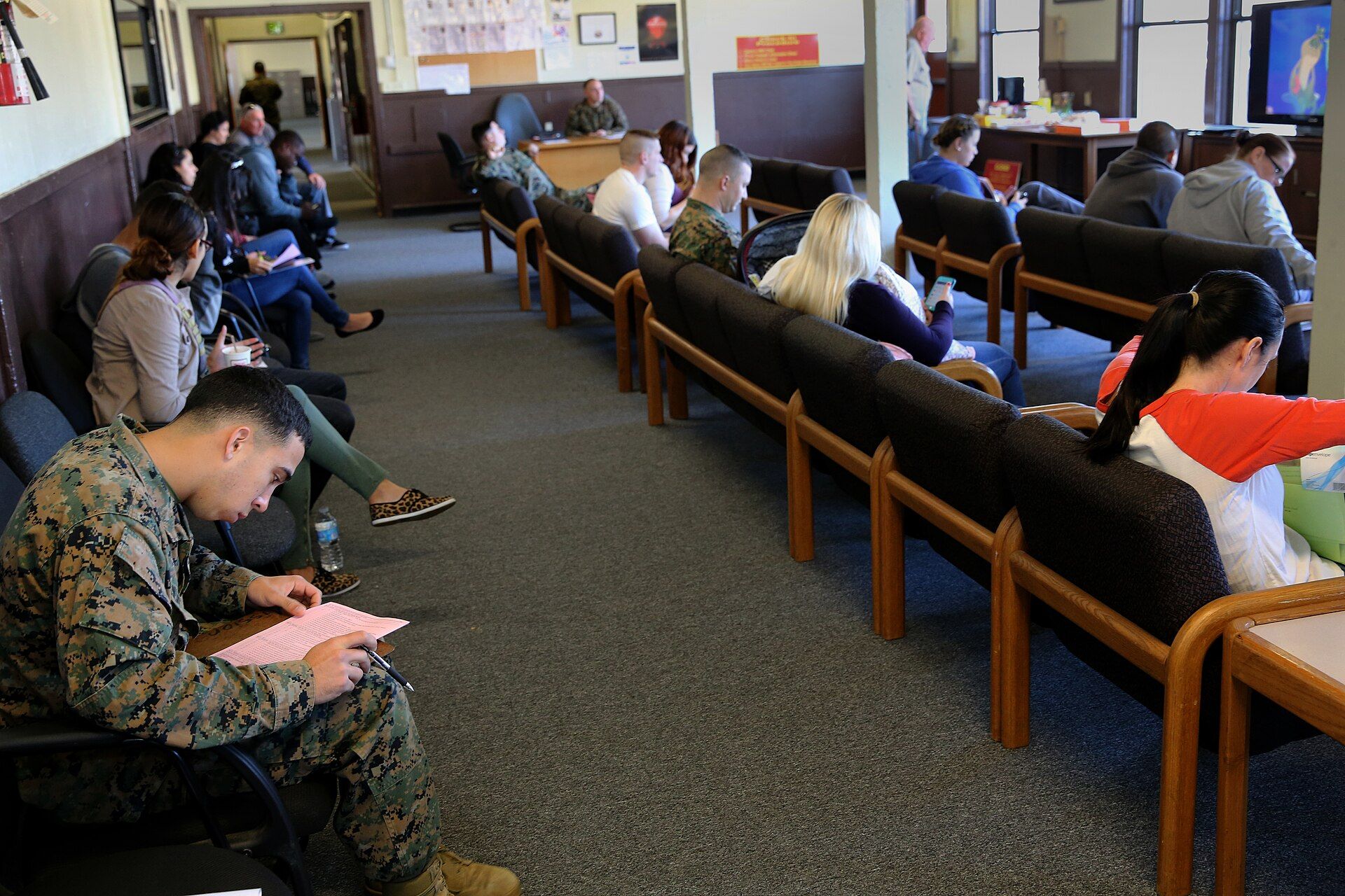 People sit on wooden benches in a waiting room, some filling out paperwork, at a tax assistance center.