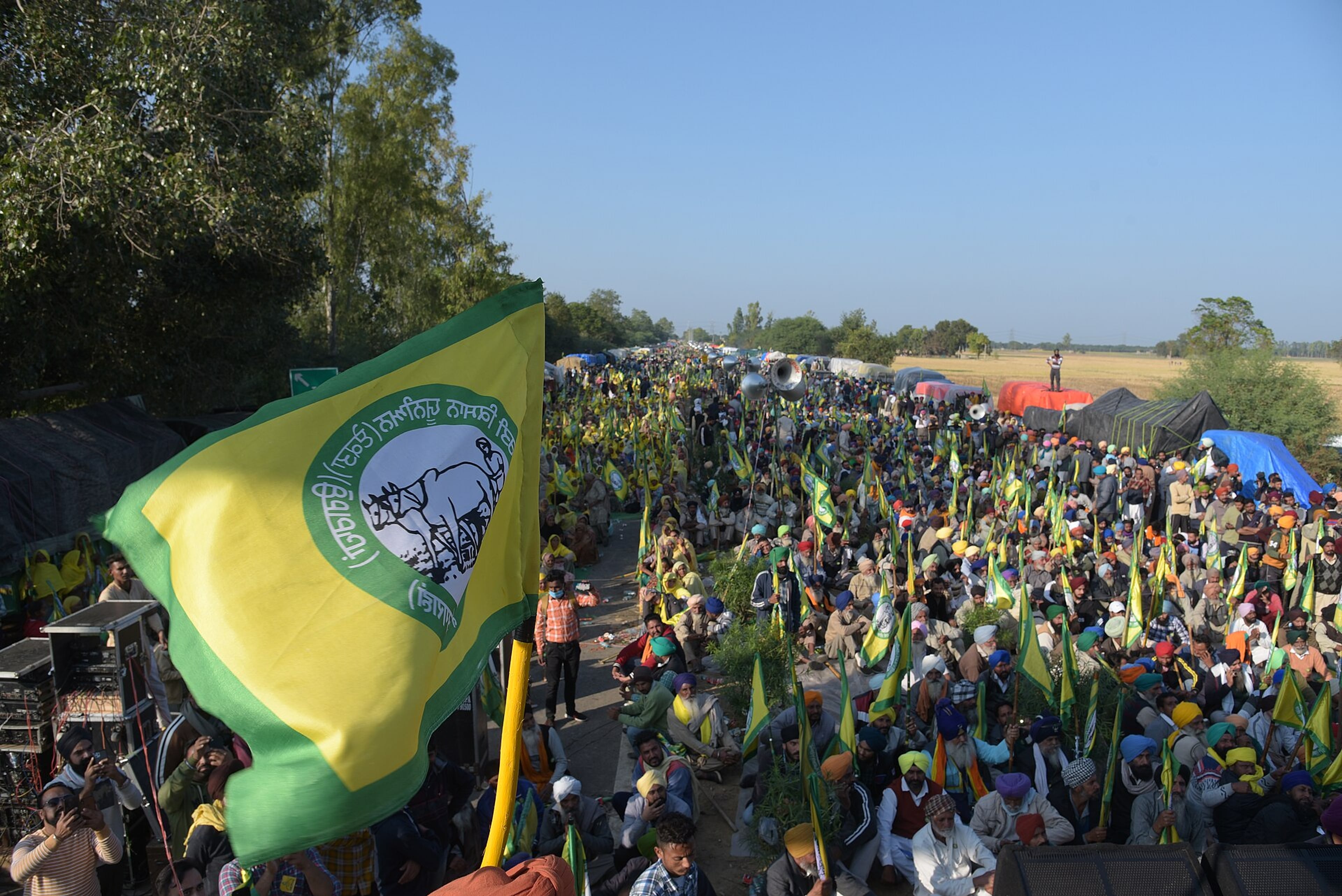 Large crowd of farmers seated at a protest site, with yellow and green union flags and tents under a clear sky.