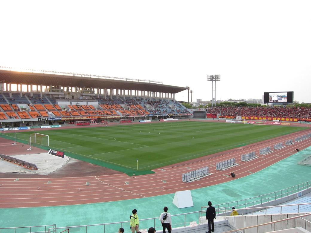 Green football pitch inside a stadium, ringed by a red running track and tiered grandstands under a pale sky, with a few people by the rail.