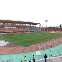 Green football pitch inside a stadium, ringed by a red running track and tiered grandstands under a pale sky, with a few people by the rail.