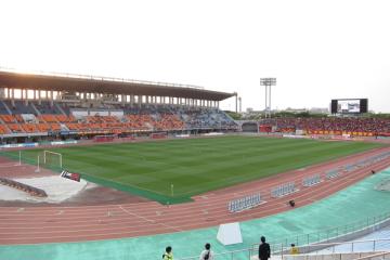 Green football pitch inside a stadium, ringed by a red running track and tiered grandstands under a pale sky, with a few people by the rail.