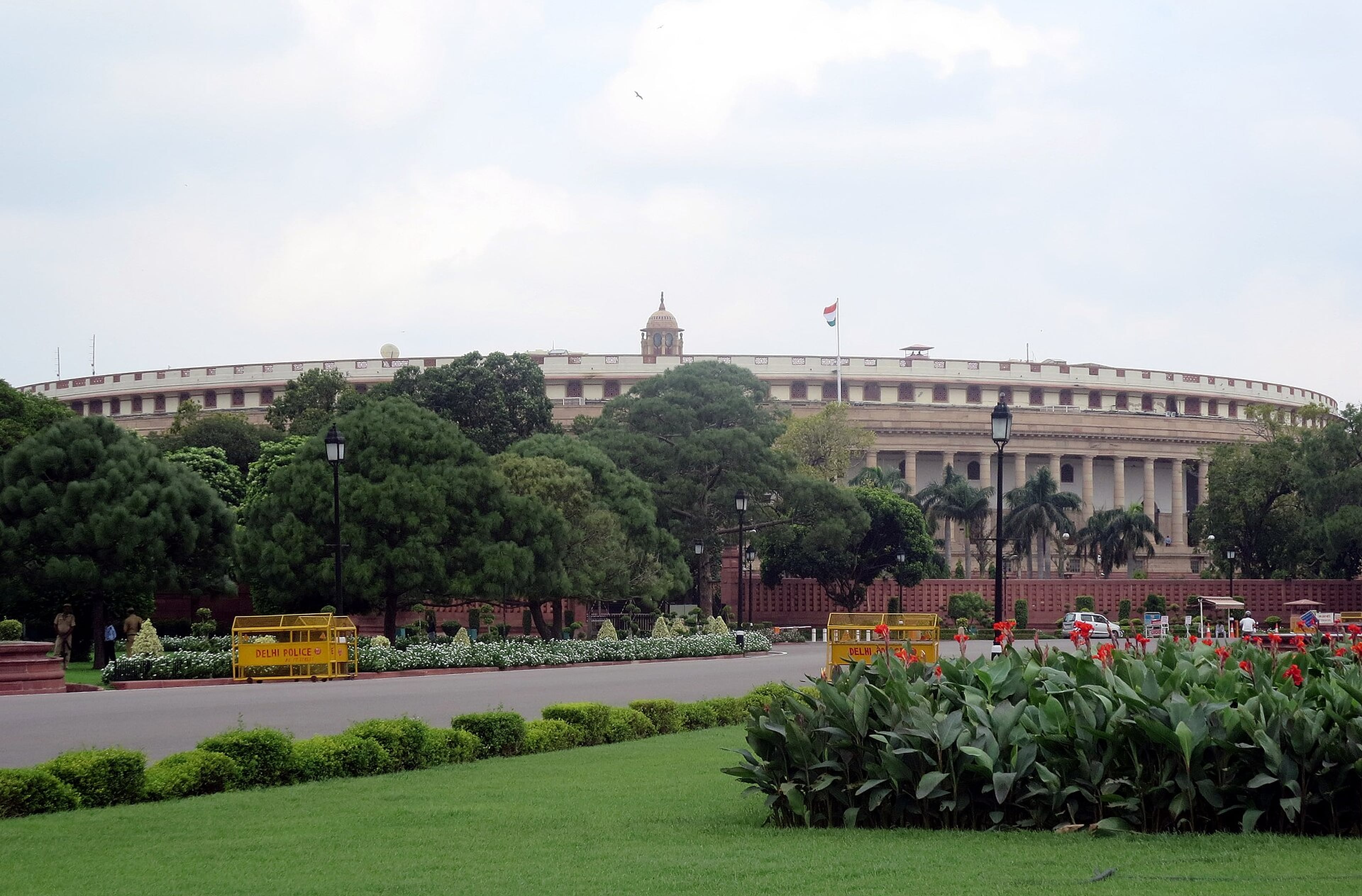 Curved, colonnaded Parliament building in New Delhi behind tall trees, with a lawn and flowerbeds in the foreground under a pale sky.