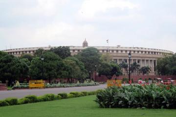 Curved, colonnaded Parliament building in New Delhi behind tall trees, with a lawn and flowerbeds in the foreground under a pale sky.