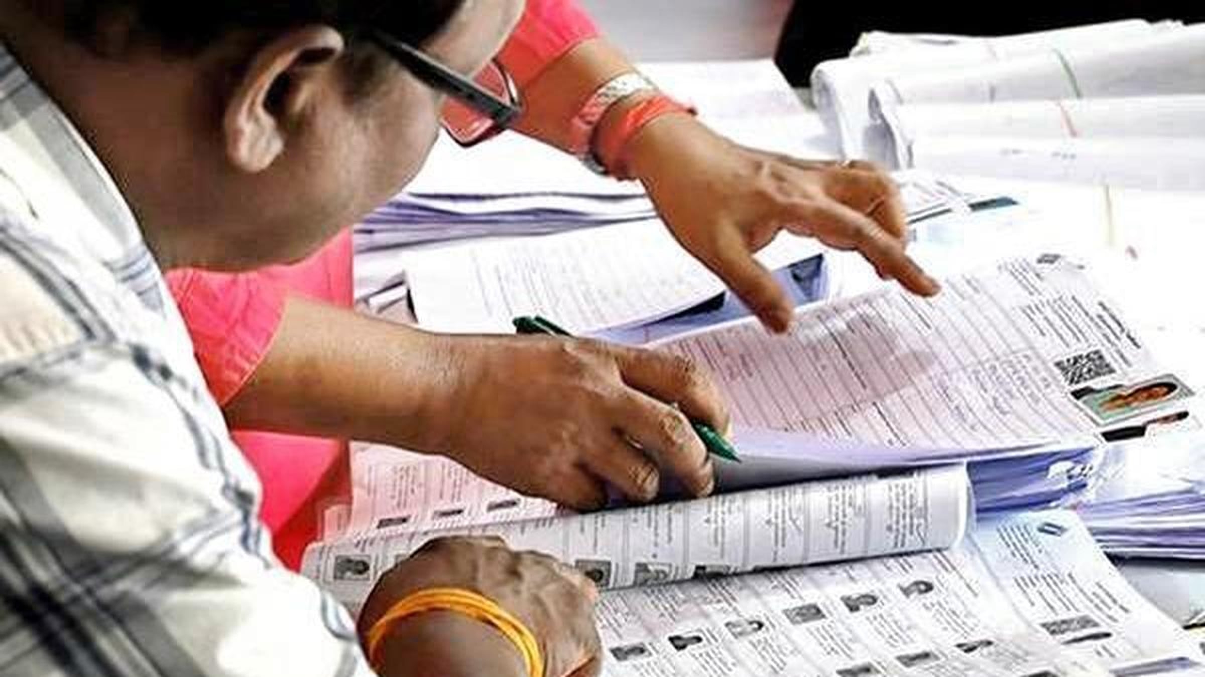 Hands and a pen on printed electoral roll pages with voter photo IDs, while a man in glasses reviews them.