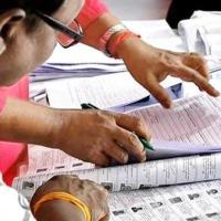 Hands and a pen on printed electoral roll pages with voter photo IDs, while a man in glasses reviews them.