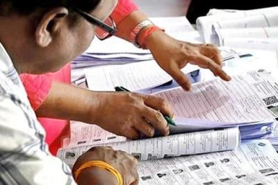 Hands and a pen on printed electoral roll pages with voter photo IDs, while a man in glasses reviews them.