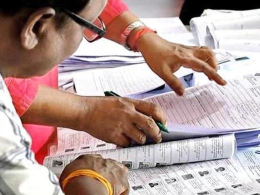 Hands and a pen on printed electoral roll pages with voter photo IDs, while a man in glasses reviews them.