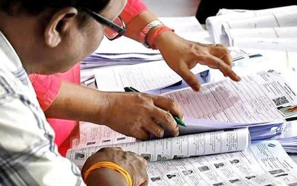 Hands and a pen on printed electoral roll pages with voter photo IDs, while a man in glasses reviews them.