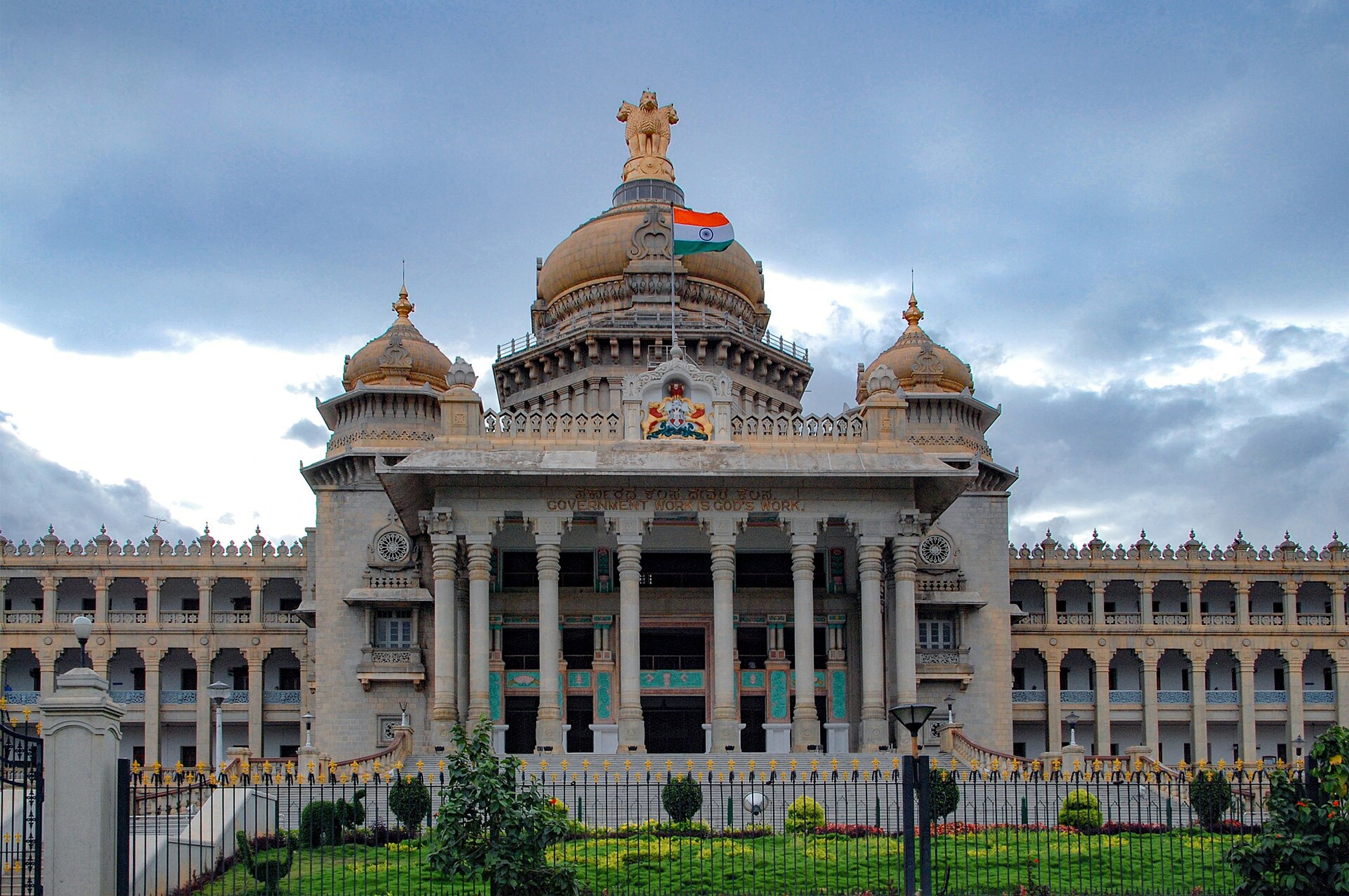 Facade of the Vidhana Soudha building in Bengaluru, with an Indian flag on its central dome under a cloudy sky