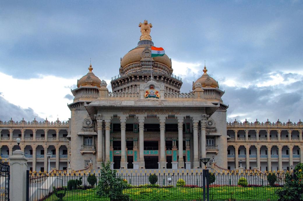 Facade of the Vidhana Soudha building in Bengaluru, with an Indian flag on its central dome under a cloudy sky